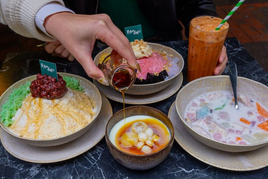 A table with several colourful desserts on it a woman is pouring a syrup over a lychee pudding