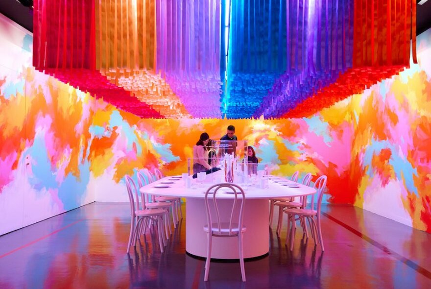People standing at a white table and chairs surrounded by colourful walls and colourful ceiling hangings above.