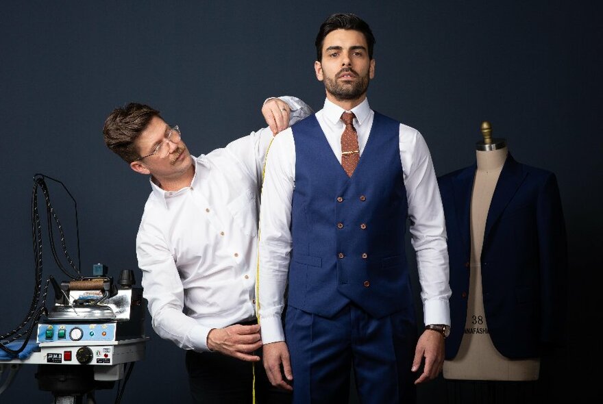 Man in blue waistcoat and trousers, looking straight ahead, being measured up for suit.