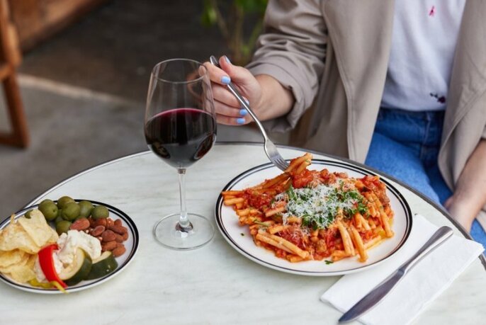 Plate of antipasti, glass of red wine, plate of pasta on marble-look table. Torso of seated patron behind with hand holding fork over pasta.