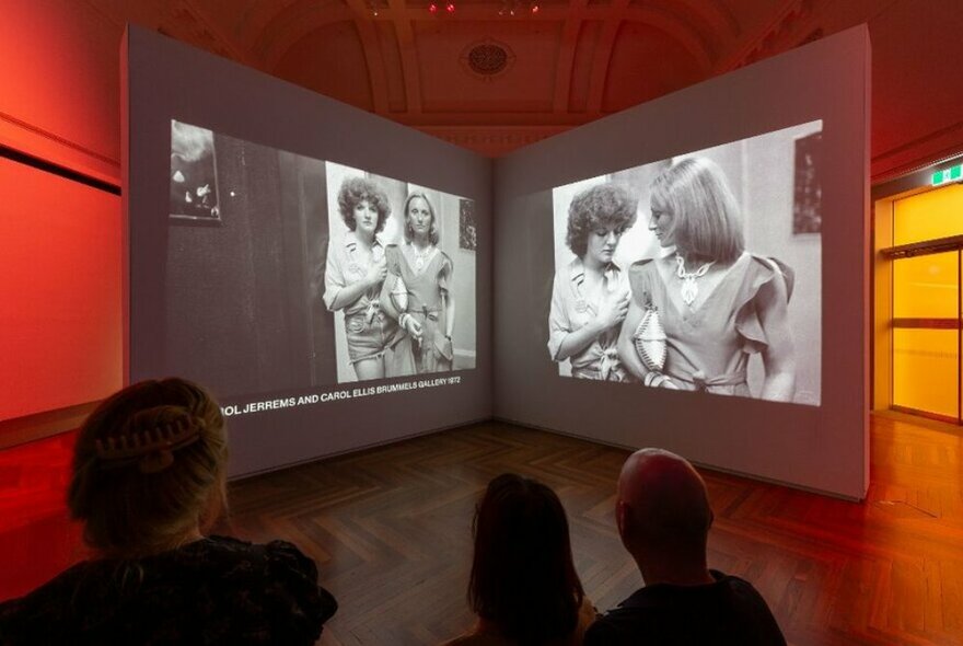 Installation view of the Rennie Ellis exhibition with two large black and white photographs projected onto a screen and people looking at them.