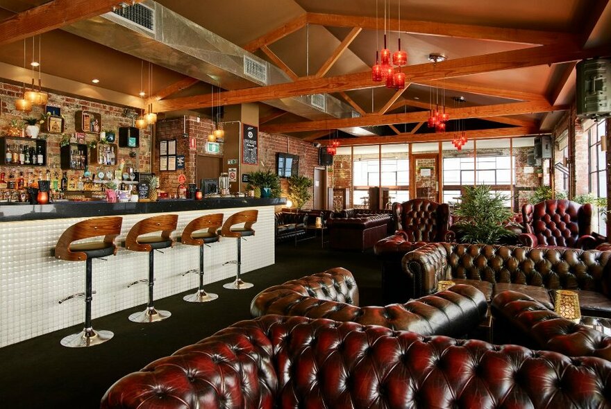 View into a bar with multiple leather couches, a central bar area with stools, large windows onto a balcony and exposed beams.