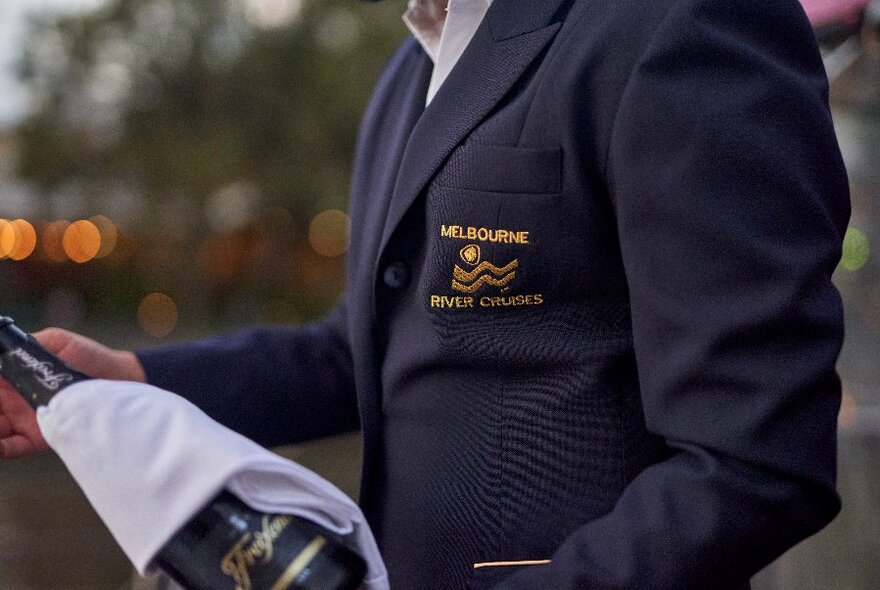 A person in a blue blazer with an emblem on the pocket about to pour champagne from a bottle with a napkin draped across it. 