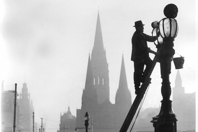 Black and white photo of lamplighter on ladder at a lamp, with buildings and spires in background.
