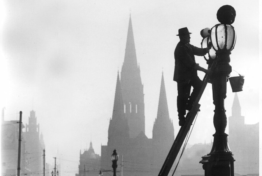 Black and white photo of lamplighter on ladder at a lamp, with buildings and spires in background.