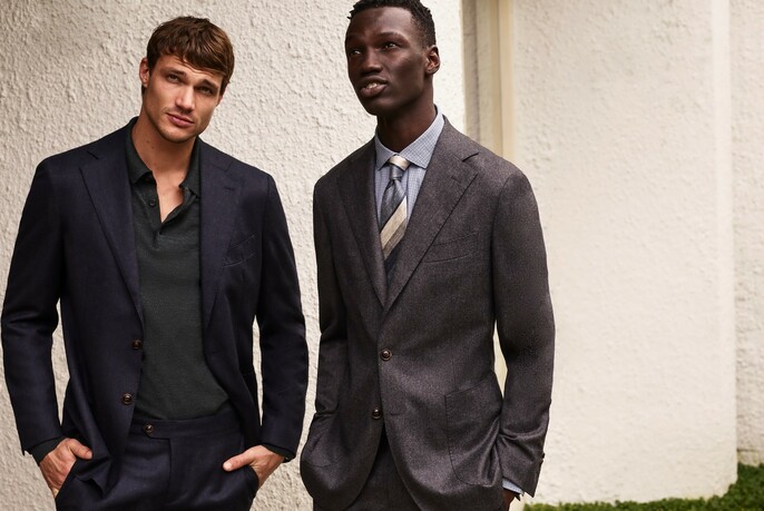 Two male models, one wearing a casual dark jacket, the other wearing a buttoned-up grey suit.
