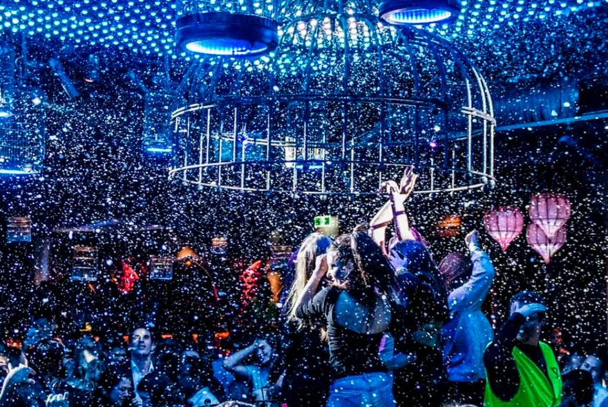 Inside a packed nightclub with people dancing under an illuminated ceiling with confetti raining down. 