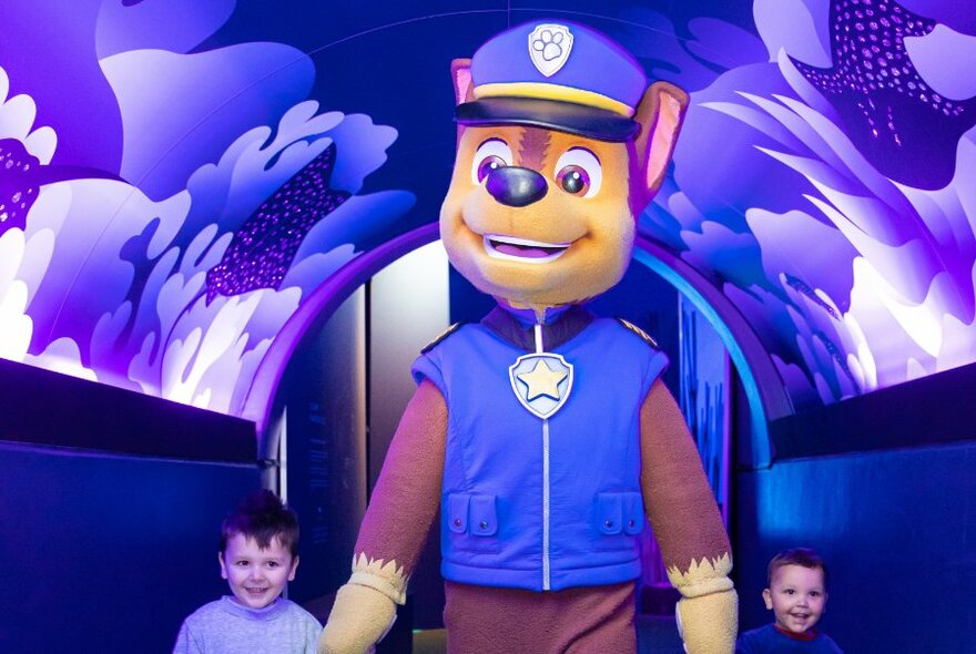 A person in a Chase costume from PAW Patrol, holding hands with two young children and walking through a tunnel with blue seaweed motifs.