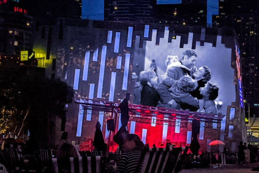 An audience seated in deckchairs at Fed Square, watching a classic Christmas film projeccted onto the big screen behind the stage.