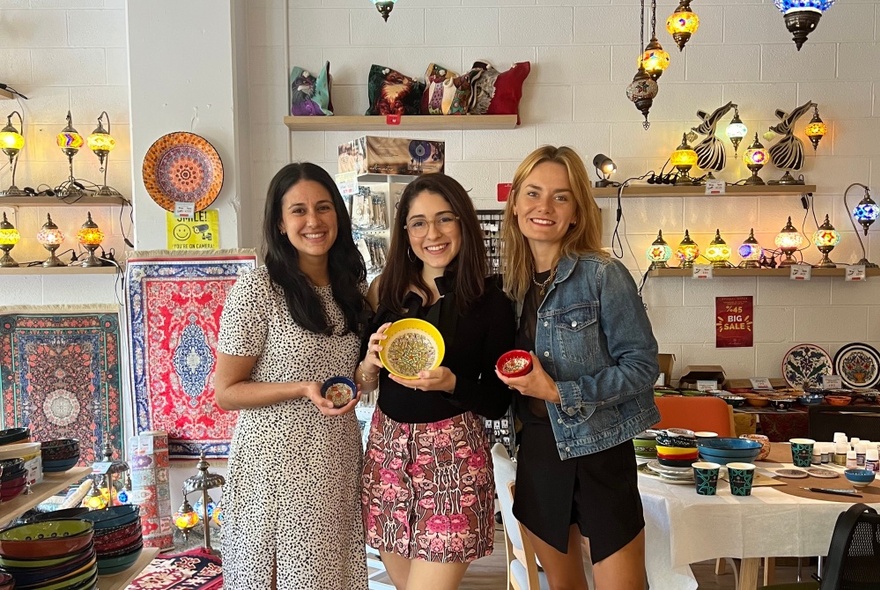 Three people standing in a shop decorated with Turkish lamps, each holding a painted bowl.