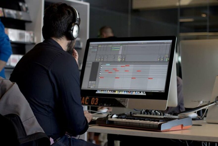 Person wearing headphones seated at a computer with piano keyboard.