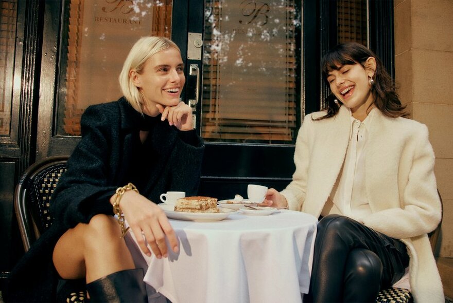 Two women smiling and modelling designer clothes, one wearing all black, the other a cream coloured coat and black pants, sitting at a cafe table outdoors.
