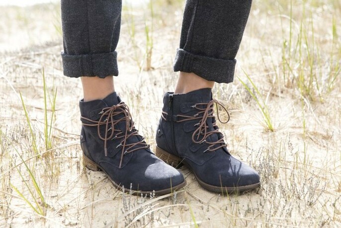 Models feet with turned-up jeans and lace-up ankle-length boots standing on grass and sand.