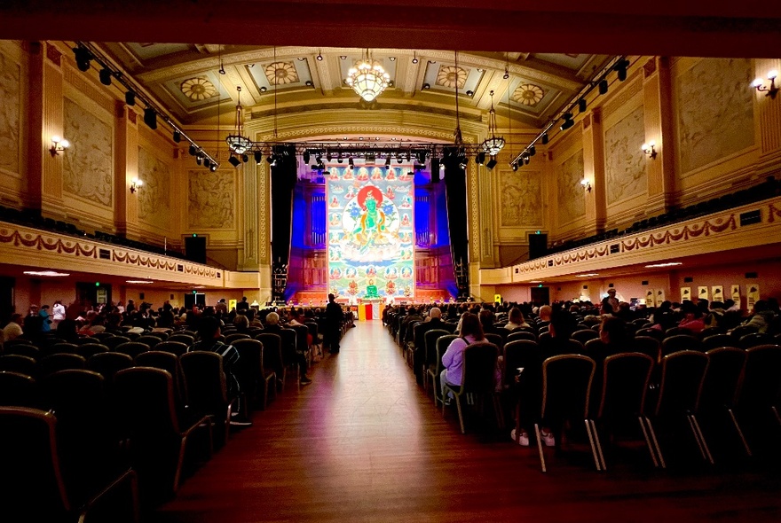 Melbourne Town Hall venue with people seated in rows looking at a huge Buddhist artwork displayed on stage.