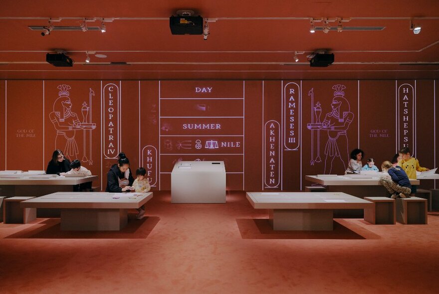 Children and families sitting at large craft tables in a spacious exhibition room, the walls decorated with ancient Egyptian drawings and hieroglyphics. 