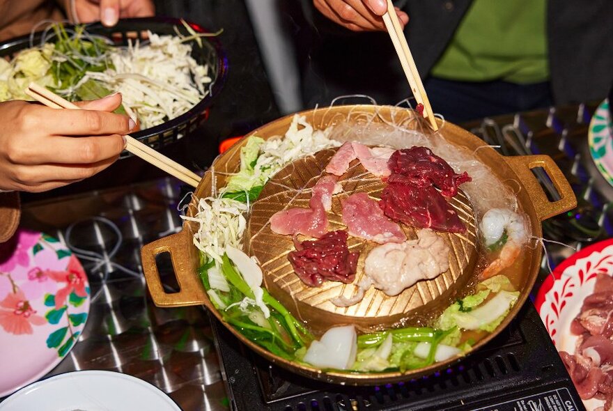 People reaching for strips of meat with chopsticks on a gold tabletop barbecue.
