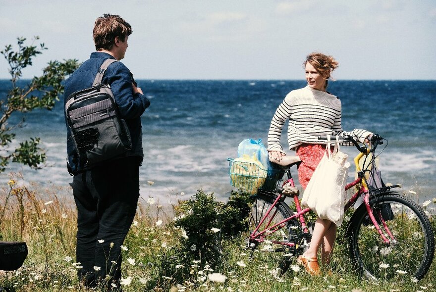 A woman on a bicycle with shopping bags stops to talk to a man with a shoulder bag in a flowery field beside the ocean. 