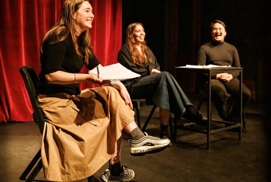 Three people on stage, smiling and laughing as they test out a new performance, red curtain in the background.