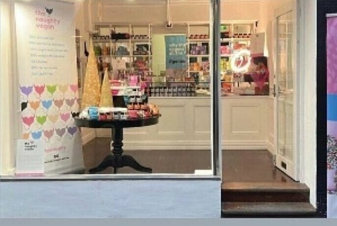 View into a tiny vegan chocolate shop with wooden floors, a table and a counter with shelves loaded with products.