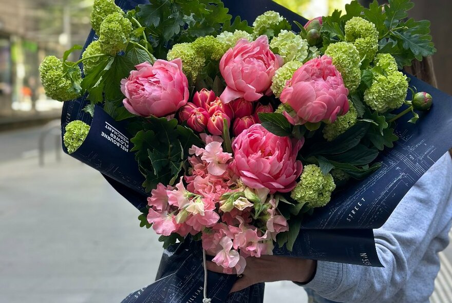 A hand holding a floral bouquet of flowers in shades of pink and green wrapped in distinctive branded dark paper.