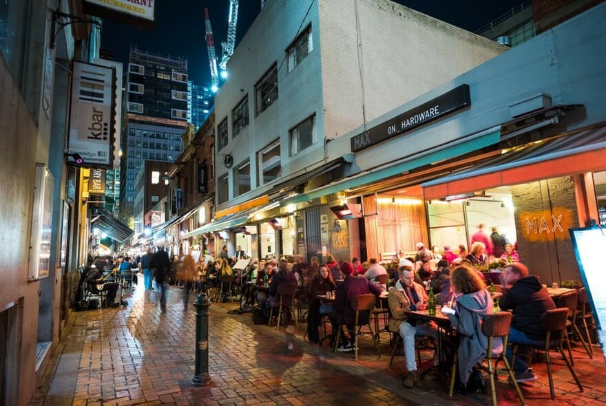 Laneway diners seated at outdoor tables on brick paving outside a restaurant at night.
