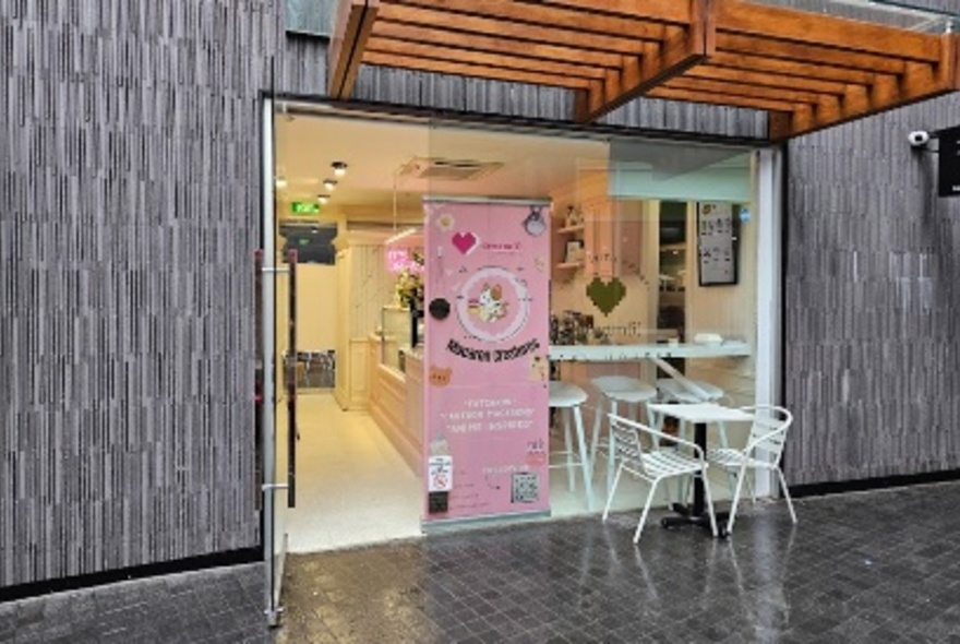 The exterior of a small store with grey tiles outside and a pink sign, with a white table set outside. 