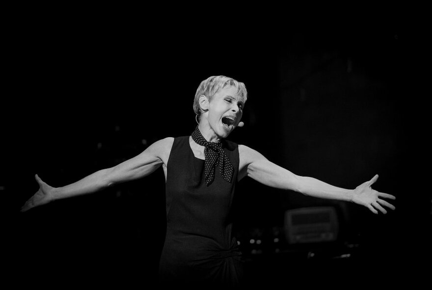 Woman with short hair and outstretched arms on a darkened stage singing expressively.