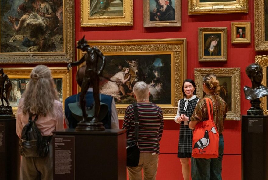 A guide leading a tour in the NGV, standing in front of a red-painted gallery wall covered in gilt-framed paintings and black sculptures.