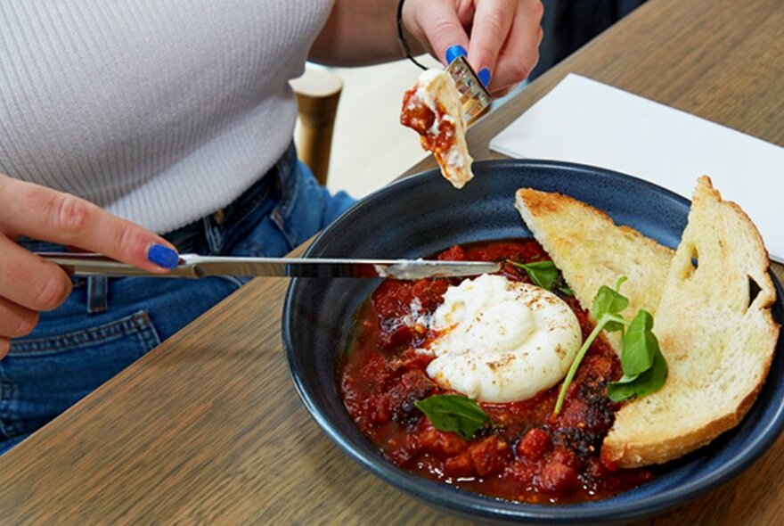 Person using cutlery to cut into a dish of baked eggs and toasted bread.