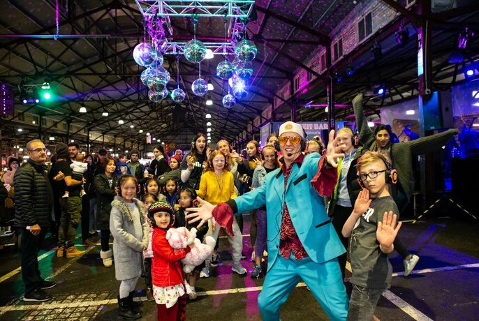 Crowd of people on a flash mob, all wearing headphones, posing in one of the aisles of the Queen Vic market sheds, with passers by watching.
