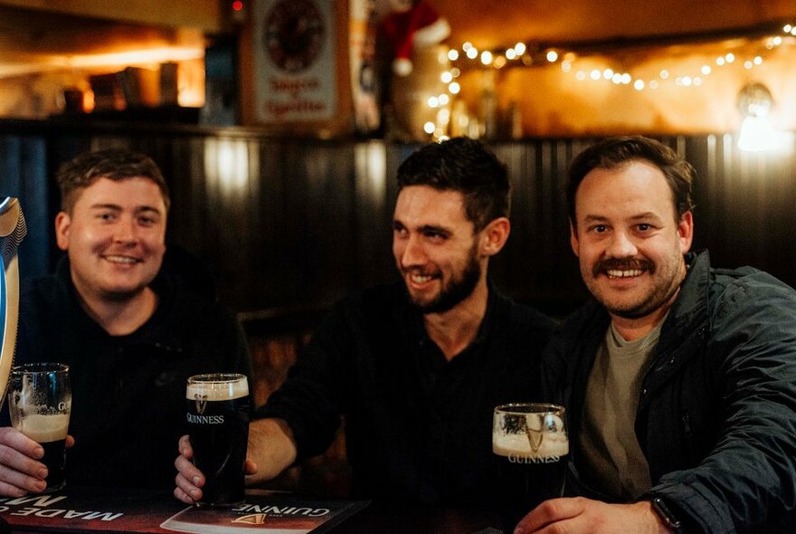Three men sitting in a dark-wood panelled pub holding glasses of Guinness.