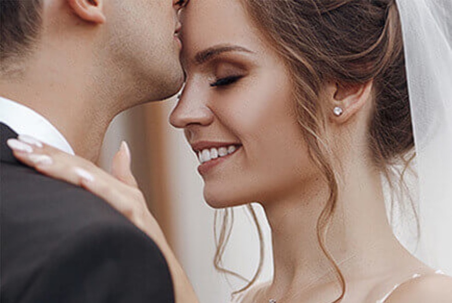 A close up of a bride and groom. The bride is smiling and wearing diamond earrings. 