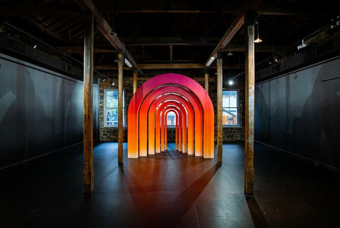 A sculpture of orange coloured cascading arches, lined up one under the other, in the middle of a gallery room of a dark floor and dark walls.