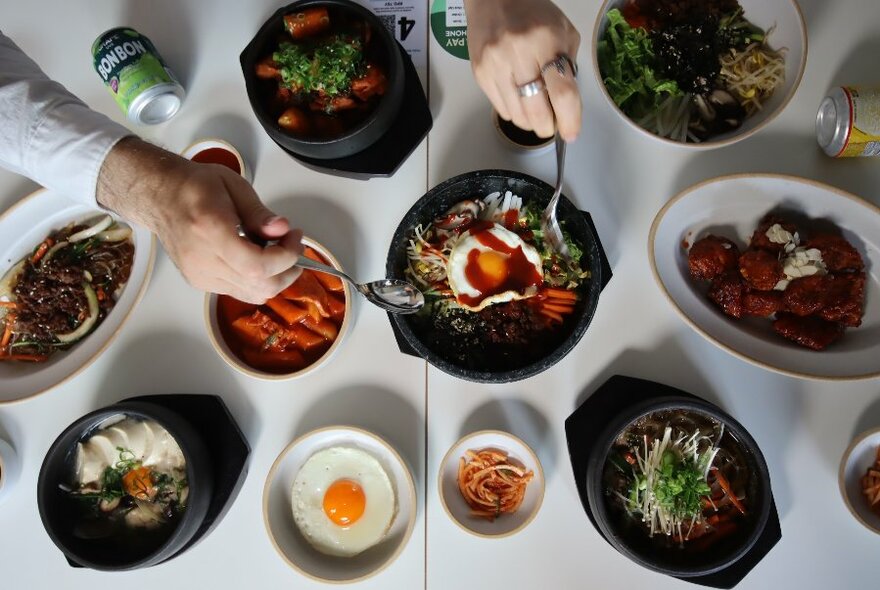 Overhead view of hands holding spoons over a range of Korean dishes on a white table, including sizzling platters, fried egg and condiments.