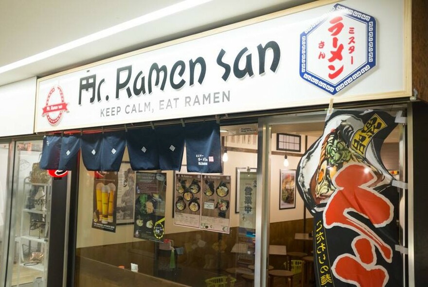 Mr Ramen San exterior with signage and script, with view into cafe seating area and food posters.