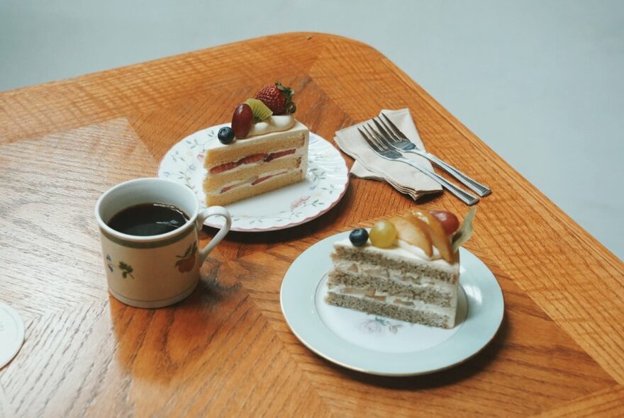 Two slices of cake on plates, with one mug of coffee to left, and forks on serviettes to right, on wooden table.