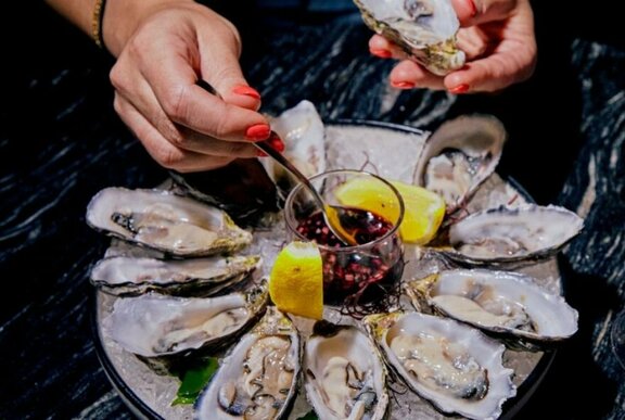 Hand hovering over a plate of shucked oysters, selecting one with a small fork and dippin it into a bowl of sauce.