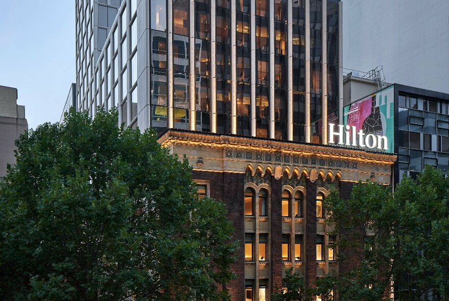 The exterior of Hilton Melbourne Little Queen Street with a large city building towering over from behind. 