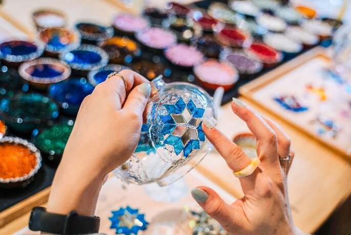 Hands positioning glass mosaic tiles onto a clear glass lamp at a craft table. 