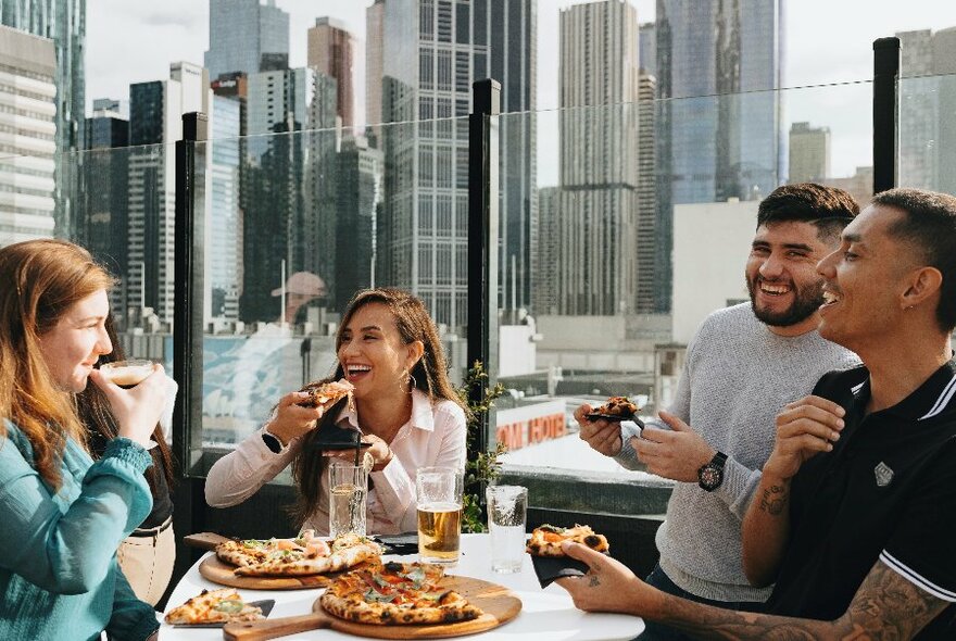 Four people seated at a rooftop table enjoying pizza and beer with city views in the background.