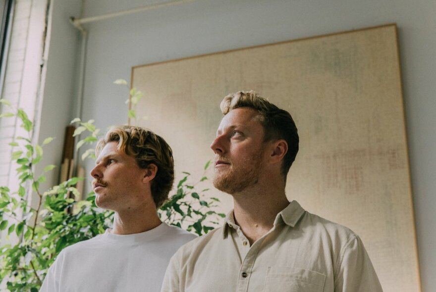 The two members of the band Hollow Coves, men in their late 20s, standing side by side and looking out to the right through a window, plants and a painting in the background behind them.