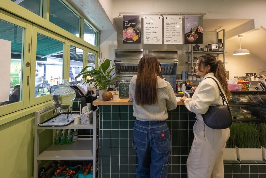 Small interior of Pandan Dessert Bar, with two patrons lined up and waiting at the counter and a view into the small kitchen and preparation area.