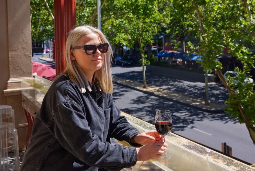 A woman drinking a glass of wine on a balcony overlooking a leafy street.