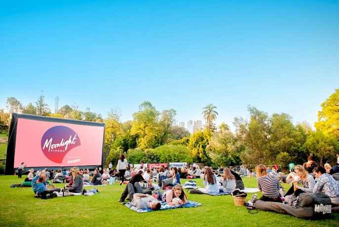 People seated on rugs on the grass in front of a big screen.