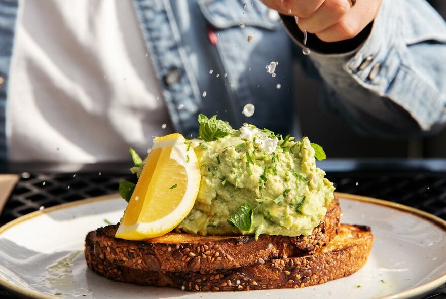 A plate of toast and smashed avocado with a slice of lemon.