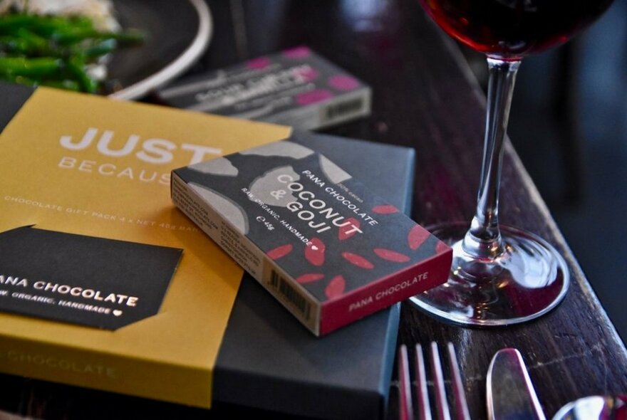 Chocolate in branded cardboard packaging resting on a dark table top, a glass of red wine to the side.  