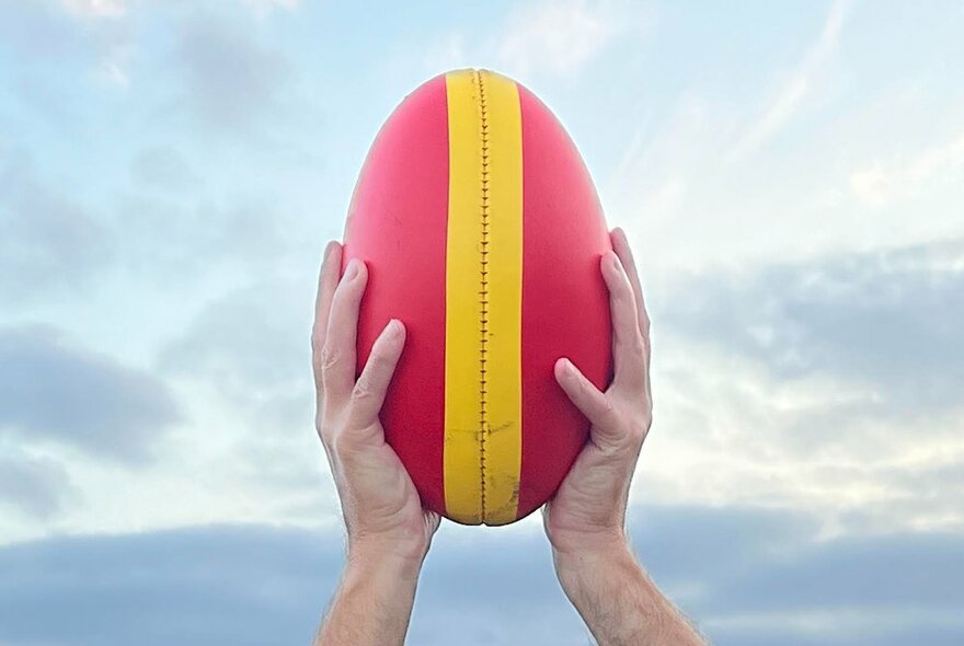 A red AFL football with a yellow stripe down the middle being held high by a pair of hands, a pale blue sky in the background.