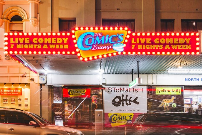 Exterior of the Comic's Lounge with brightly lit neon signage and cars driving by on the street. 