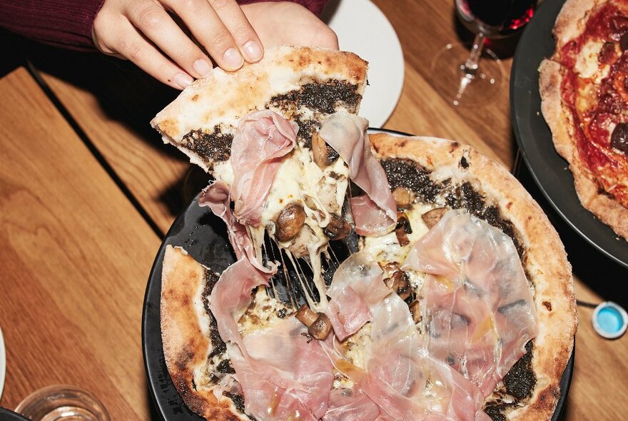 Hands pulling a slice of pizza covered in prosciutto. 