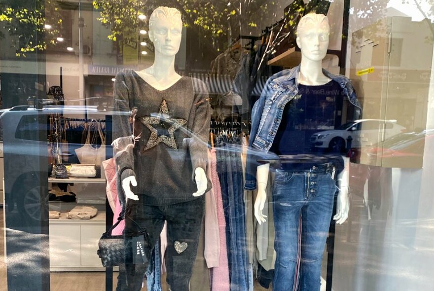 A boutique shop window with reflected streetscape with cars, the two mannequins wearing casual trousers and jumpers.